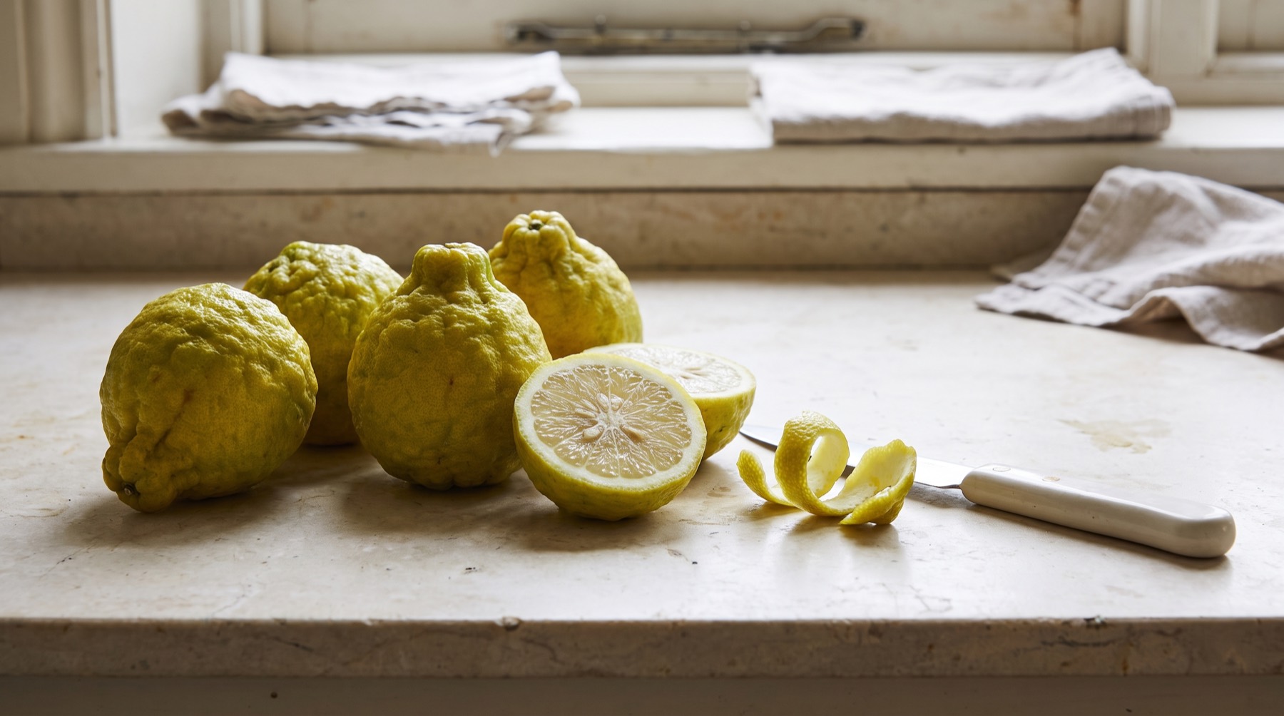 Whole bergamot fruits with a knobbly yellow green rind, one cut in half, and a strip of peel on a stone worktop